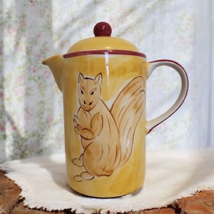 May include: A yellow ceramic pitcher with a red lid and a painted squirrel design. The squirrel is brown and has a bushy tail.