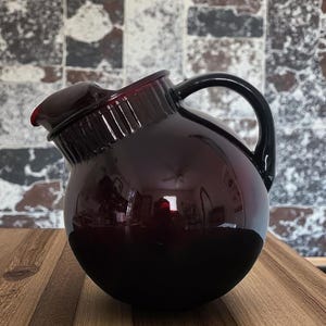 May include: A dark red glass pitcher with a black handle and a curved spout. The pitcher has a round body and a glossy finish. The pitcher is sitting on a wooden surface.