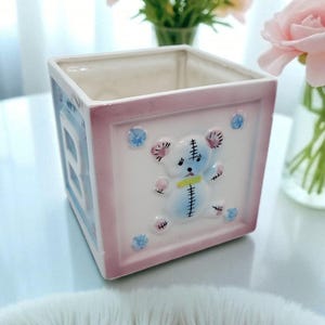 May include: A square ceramic planter with a baby block design. The planter features a light pink border and a white center with a blue teddy bear illustration. Blue and pink floral accents decorate the sides.