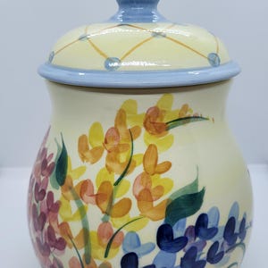 May include: A ceramic jar with a lid, featuring a floral design with yellow, orange, pink, blue, and green flowers. The lid is light blue with a yellow and white pattern.