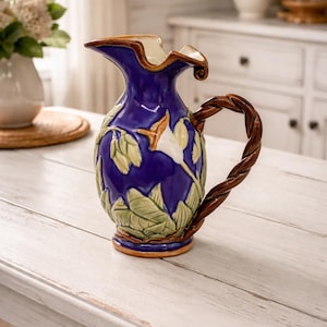 May include: A decorative blue ceramic pitcher with a brown twisted handle. The pitcher features a floral design with green leaves and white flowers. The rim is cream-colored, and the base is brown.