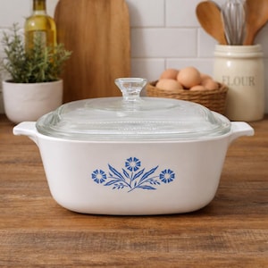 Puede incluir: Cazuela blanca con tapa de cristal transparente. El plato tiene un diseño floral azul en la parte delantera y asas blancas a cada lado. El plato está sobre una superficie de madera.