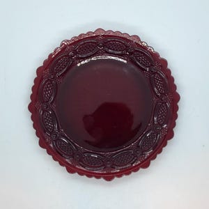 May include: A round, ruby red glass plate with a scalloped edge. The plate features a detailed embossed pattern around the rim, with a smooth, glossy center. The plate is likely vintage and suitable for serving or display.