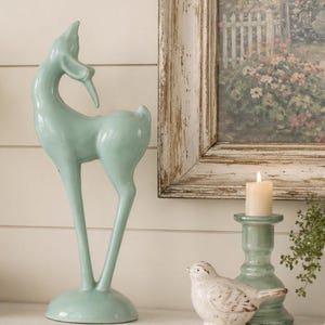 May include: A smooth, light mint green ceramic deer figurine standing on a circular base, accompanied by a small white speckled ceramic bird figurine and a light mint green candle holder with a single lit white pillar candle.