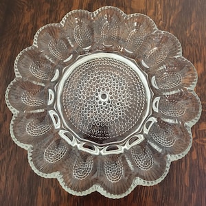 May include: Clear glass serving dish with a scalloped edge and a central circular design. The dish features a pattern of small, raised circles in the center and oval-shaped indentations around the rim. The glass is transparent, allowing the dark wood surface beneath to show through.