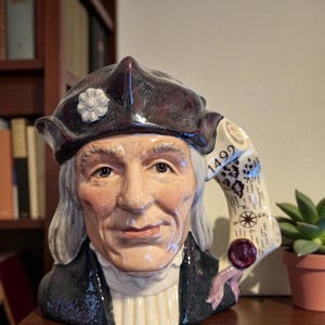 May include: A ceramic mug shaped like a man's head, featuring a dark hat with a white flower, long white hair, and a black coat. The mug's handle is a scroll with the year 1492 and a map design. The mug is on a wooden surface.