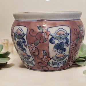 May include: A small, round, ceramic planter with a pink, white, and blue floral design. The planter has a slightly flared rim and a rounded base.
