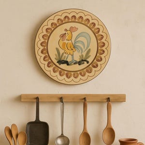 May include: A round, decorative plate featuring a yellow rooster with a blue tail and red comb. The plate has a scalloped edge with a brown leaf pattern. Below, a wooden rack holds various kitchen utensils, including spoons and a small pan.