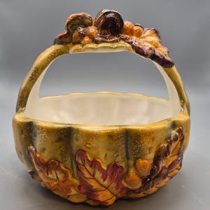 May include: A ceramic basket with a glossy finish, featuring a sculpted pumpkin shape with a handle. The exterior is decorated with raised, hand-painted autumn leaves and acorns in shades of orange, brown, and yellow, with a smooth cream interior.