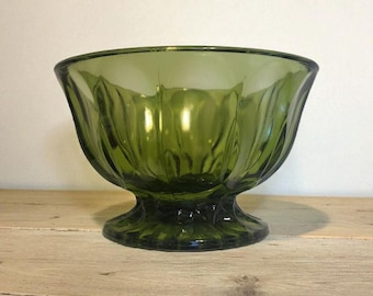 May include: A vintage green glass dessert bowl with a pedestal base. The bowl has a fluted design around the sides and a rounded rim. The bowl is sitting on a wooden surface.