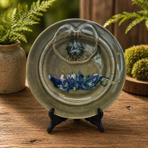 May include: A ceramic decorative plate with a frog sculpture in the center. The plate is olive green with blue and green abstract paint strokes. It is displayed on a black stand, showcasing the handcrafted artistry.