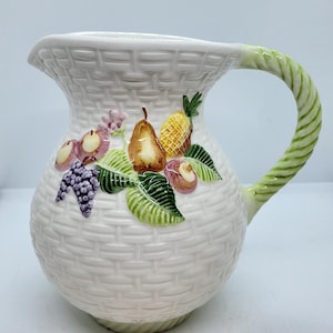 May include: White ceramic pitcher with a woven basket design. The pitcher features a green, twisted handle and is decorated with colorful, raised fruit including grapes, pears, and a pineapple. A decorative piece for serving beverages.