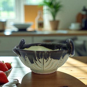 May include: A ceramic bowl with a leaf design, featuring a dark brown rim and handles. The bowl has a light cream interior and is set on a wooden surface. Strawberries are visible in a small bowl to the left.