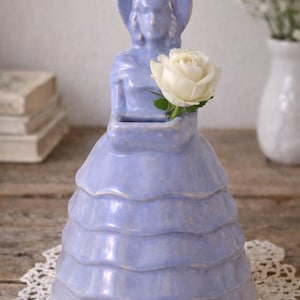 May include: A lavender-colored ceramic vase shaped like a woman in a dress, holding a white rose. The vase has a hat and a tiered skirt design. The vase is on a white lace doily.
