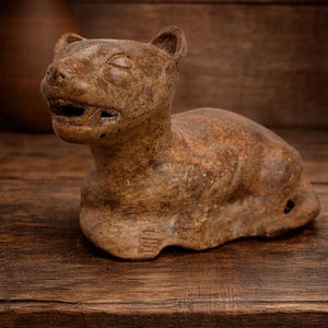 May include: A brown, hand-carved sculpture of a feline animal, possibly a cougar or jaguar, with its mouth slightly open. The sculpture is made of a stone-like material and rests on a wooden surface. The piece has a rustic, aged appearance.