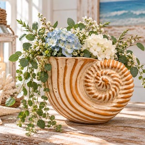 May include: A ceramic seashell-shaped planter with a cream-colored base and brown ribbed detailing, filled with an arrangement of blue and white hydrangea flowers, small white blossoms, and green eucalyptus-style foliage.