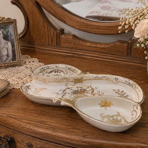 May include: A white ceramic divided serving dish featuring an elongated, asymmetrical shape with three distinct sections. The dish is decorated with intricate gold filigree, floral patterns, and scrollwork accents along the interior rims and base.