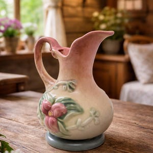 May include: A vintage ceramic pitcher with a pink and cream color scheme. The pitcher features a handle and a spout, with a floral design in pink and green. It sits on a light blue base, placed on a wooden table.