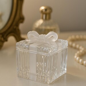 May include: A clear glass box with a lid, adorned with a white ribbon and bow. The box has vertical ribbed detailing and sits on a light-colored surface. A gold-topped perfume bottle and a string of pearls are in the background.