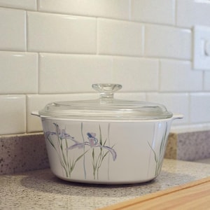 May include: White casserole dish with a clear glass lid. The dish features a floral design of purple irises and green stems. The dish is on a speckled countertop.