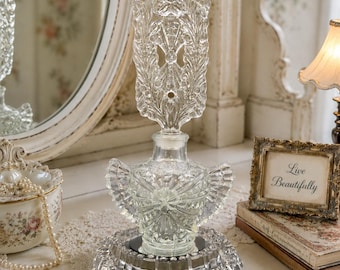 May include: A clear glass perfume bottle with a tall, ornate, leaf-patterned stopper and a wide, faceted base. The bottle features a textured, wing-like design on its body and sits atop a circular, reflective glass stand with a scalloped edge.