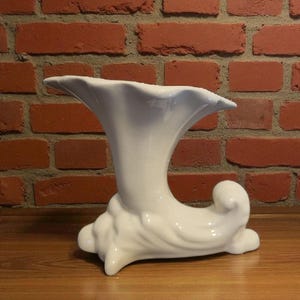 May include: A white ceramic vase with a flared, ruffled top and a curved, sculptural base. The vase has a smooth, glossy finish and sits on a wooden surface against a red brick wall.