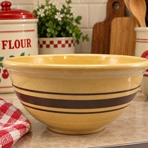 May include: A round, yellow ceramic mixing bowl featuring two horizontal dark brown stripes encircling the exterior. The bowl has a smooth, glossy finish with a wide, rounded rim and a tapered base, designed for kitchen use and food preparation.