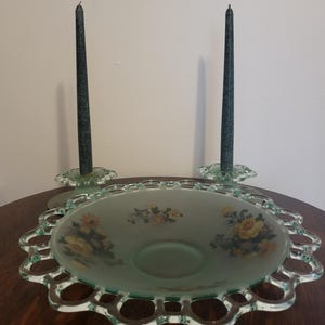May include: A vintage glass centerpiece with a scalloped edge, featuring a frosted green bowl with floral designs and two dark green taper candles. The candle holders are also green glass.