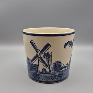 May include: A cylindrical ceramic planter with a cream-colored base and a hand-painted blue design featuring a traditional windmill, a small house, and trees. The rim of the planter is accented with a thin, solid blue line around the top edge.