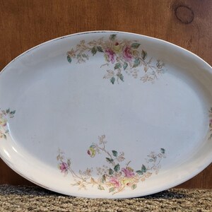 May include: An oval, off-white serving platter with a floral design. The edges of the platter are slightly worn. The floral pattern features pink, yellow, and green flowers and leaves. The platter is likely vintage and suitable for serving food.