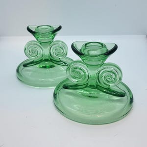 May include: Two green glass candle holders. Each has a circular base, a decorative swirl design, and a flared top to hold a candle. The glass is a translucent green color.