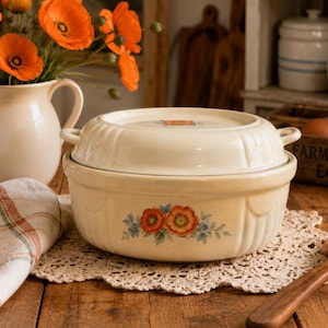 May include: A cream-colored, lidded casserole dish with floral accents. The dish sits on a lace doily, next to a wooden spoon. A white pitcher with orange poppies and a plaid cloth are also in the image.