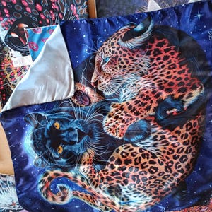 Puede incluir: Un cojín decorativo cuadrado azul y negro con un leopardo y una pantera. El leopardo tiene manchas naranjas y negras, y la pantera es negra. El fondo es un cielo estrellado. El cojín tiene un forro plateado.