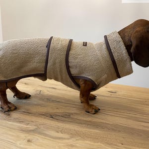 Op de afbeelding: Een beige hondenbadjas met donkerbruine randen gedragen door een bruine teckel. De badjas heeft een vierkant logo op de rug en is gemaakt van een zacht, absorberend materiaal. De hond staat op een houten oppervlak.