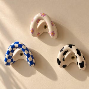 May include: Three arched wall hooks made of ceramic. One hook features a white base with purple and yellow daisy patterns, one has a blue and white checkerboard pattern, and one has a white base with black cow-print spots. Each hook has a central screw.
