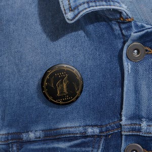 May include: A black button badge featuring gold text and a gold illustration of a staircase. The text reads "Every Step is Progress" in a circular design. The badge is set against a blue denim jacket.