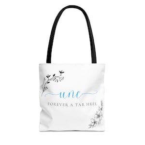 Könnte beinhalten: Weiße Tragetasche mit schwarzen Henkeln. Auf der Tasche steht das Wort "unc" in blauer Schrift, darunter der Satz "FOREVER A TAR HEEL". Florale Akzente in Schwarz befinden sich auf der linken und rechten Seite der Tasche.