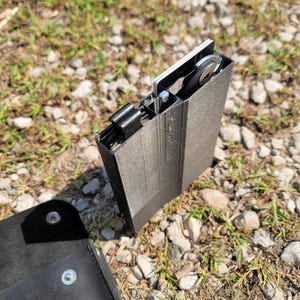 Pu&ograve; includere: Un porta carte nero stampato in 3D con una carta di metallo argentata all'interno. Il porta carte &egrave; appoggiato su un letto di piccole pietre e erba.