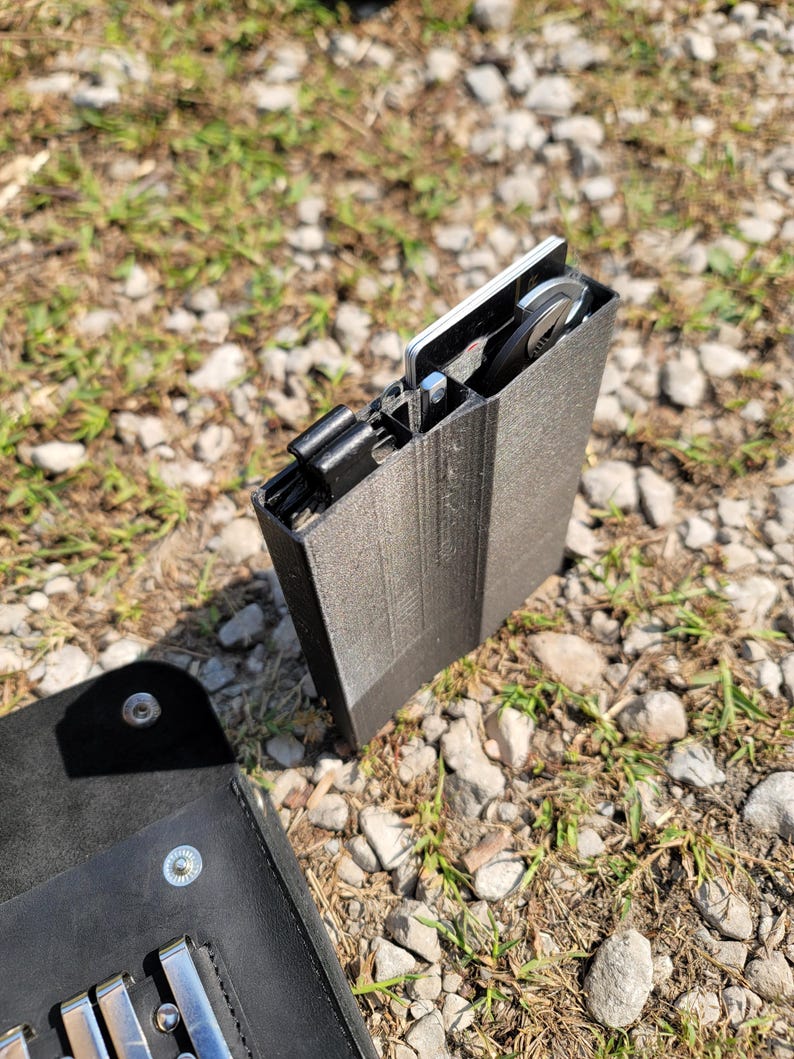 Pu&ograve; includere: Un porta carte nero stampato in 3D con una carta di metallo argentata all'interno. Il porta carte &egrave; appoggiato su un letto di piccole pietre e erba.