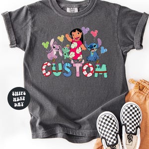 A gray t-shirt with a colorful graphic design featuring characters from the Disney movie Lilo & Stitch, along with the word "CUSTOM" written on it. The t-shirt is placed on a white surface, and there are a pair of black and white checkered shoes visible next to it.