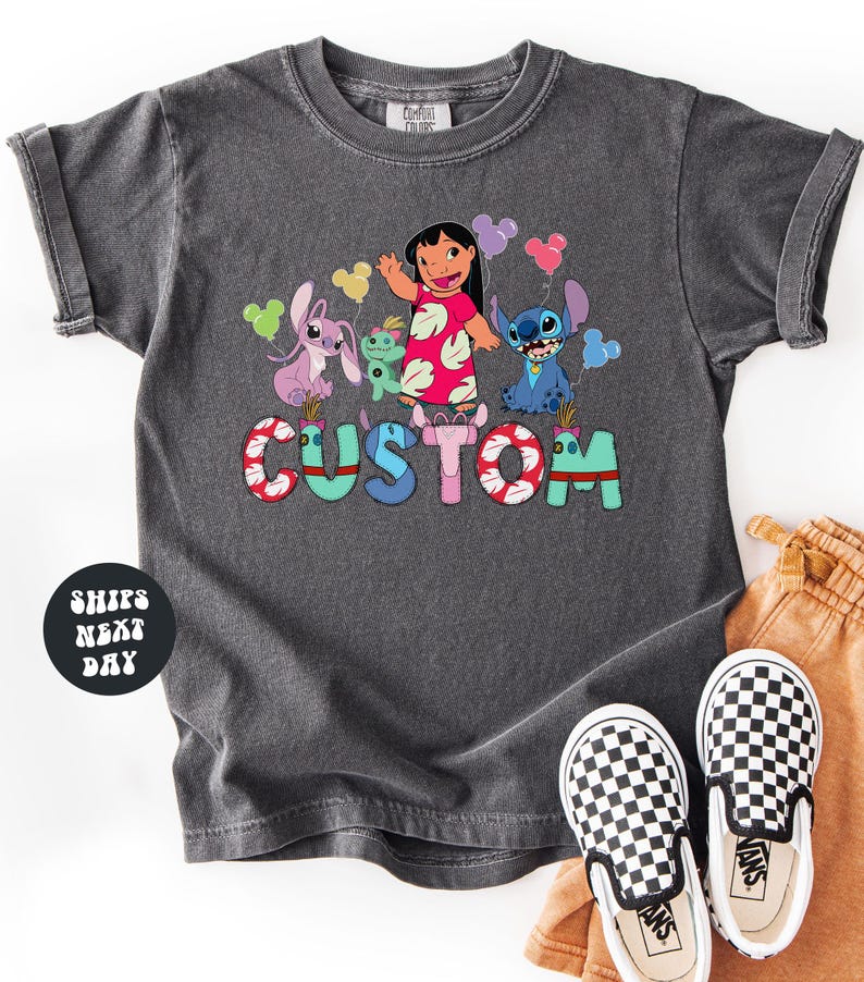 A gray t-shirt with a colorful graphic design featuring characters from the Disney movie Lilo & Stitch, along with the word "CUSTOM" written on it. The t-shirt is placed on a white surface, and there are a pair of black and white checkered shoes visible next to it.