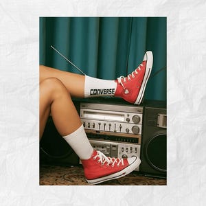 May include: Red high-top Converse sneakers with white laces and a white sock with the word "CONVERSE" printed on it. The sneakers are worn by a person sitting on a vintage stereo system.