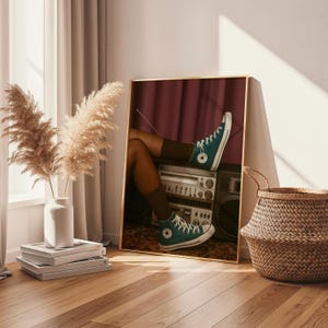 Könnte beinhalten: Ein gerahmter Druck mit tealfarbenen High-Top-Sneakern mit weißen Schnürsenkeln, die auf einem Vintage-Boombox liegen. Das Bild hat einen goldenen Rahmen und einen burgunderfarbenen Hintergrund. Die Szene umfasst einen geflochtenen Korb, Pampasgras und einen Stapel Bücher.