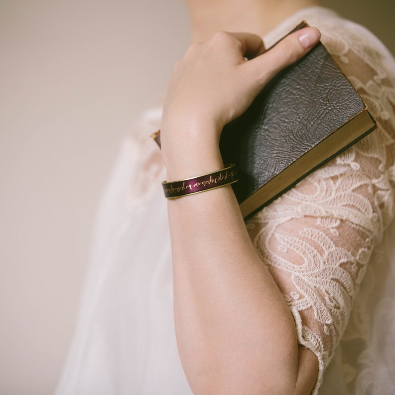 May include: A woman's wrist is adorned with a purple and gold bracelet with the words "understand up overextended" engraved on it. She is holding a brown leather-bound book.