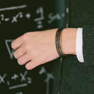 May include: A gold-toned metal bracelet with black engraved numbers. The bracelet is worn on a person's wrist.