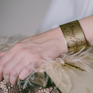 May include: Gold metal bracelet with a red inscription that reads "But I'm Damned If I Know". The bracelet is on a person's wrist.
