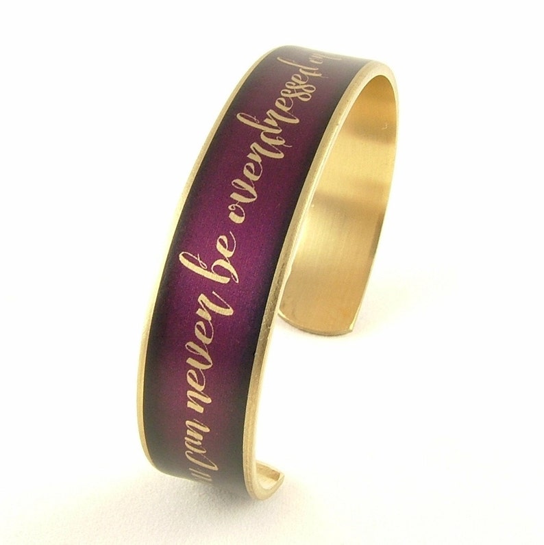 May include: A gold-toned metal bracelet with a purple band and the inscription "You were never be overdressed" in gold lettering.
