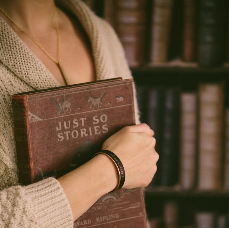 May include: A woman wearing a beige cardigan holds a vintage book titled "Just So Stories" by Rudyard Kipling. She is wearing a black leather bracelet with gold accents.