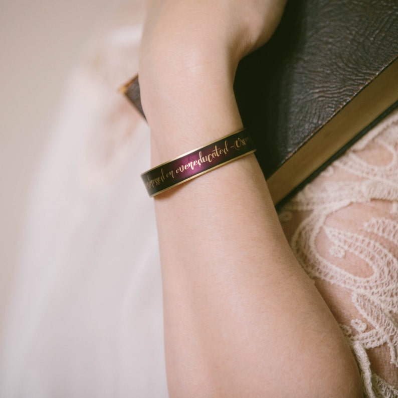 May include: A purple and gold bracelet with the inscription "Never underestimate the power of a well-educated - Ox"