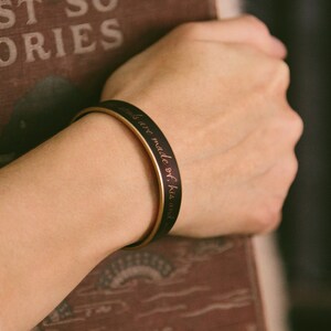 May include: A black and gold bangle bracelet with the inscription "Stories are made of his and her..." in a cursive font.
